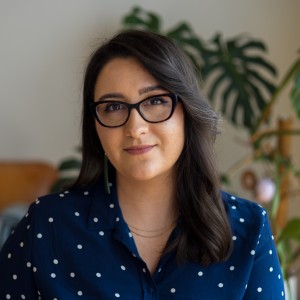 Kera Sherwood-O'Regan, a Māori and Pākehā woman with olive skin, and dark brown and grey hair and brown eyes is looking confidently at the camera. She is wearing brown vision glasses, long pounamu (greenstone) earrings, a fine gold necklace, and a navy bl