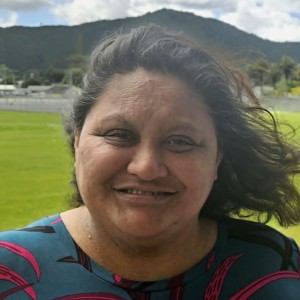 A head and shoulder photo of a Māori woman with a green field and hills in the background. Her black hair is blowing in the wind, and she is smiling.